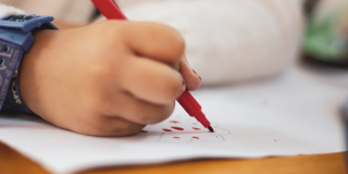 Primer plano de la mano de un niños escribiendo en un papel con un marcador rojo.