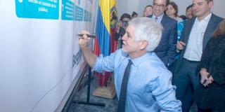 Alcalde Peñalosa viste camisa y corbata azul y se agacha para firmar un tablero.