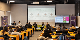 Imagen de salón donde tres personas exponen cifras con una gran imagen con los resultados, de frente un auditorio presta atención