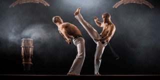 Imagen de dos hombres de pantalon blanco y sin camisa practicando capoeira