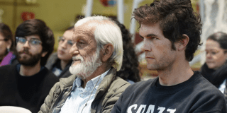 Imagen de varios hombres de diferentes edades sentados prestando atención hacía un escenario.