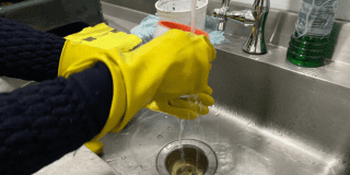 Imagen de unas manos que usan unos guantes amarillos lavando en un lavaplatos gris un vaso de cristal