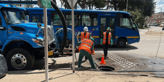 Imagen de tres técnicos de la Empresa de Acueducto realizando mantenimiento en un tubo del acueducto de una calle de la ciudad, al fondo se ven un carrotanque y varios buses del SITP.