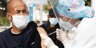 Imagen de una mujer con vestido se seguridad aplicando una vacuna a un hombre que está sentada en una silla