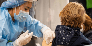 Imagen de una mujer con vestido se seguridad aplicando una vacuna a un mujer que está sentada en una silla