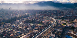 Panorámica aérea de Bogotá con trazado urbano, corredor vial elevado y cerros al fondo bajo neblina