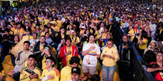 Multitud numerosa reunida en fanzone del Parque El Tunal observa partido de la Selección Colombia en pantallas gigantes; asistentes visten camisetas amarillas, sostienen banderas, teléfonos móviles y aplauden de pie en jornada nocturna.