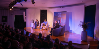 Panel de conversación en auditorio con cuatro personas sentadas en el escenario frente a público asistente, iluminación azul y morada, plantas decorativas y banner al fondo.