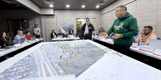 Autoridades distritales participan en simulación de manejo de emergencias alrededor de una mesa con mapas a gran escala, fichas de ubicación y material de planificación.