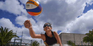 Jugadora de voleibol playa golpea un balón Mikasa durante un partido sobre arena, con gafas deportivas, malla de voleibol y palmeras visibles bajo un cielo parcialmente nublado.