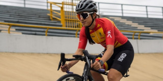 Paradeportista del Equipo Bogotá entrena ciclismo de pista en velódromo, usando casco y uniforme institucional rojo y amarillo, mientras pedalea sobre superficie inclinada con graderías metálicas al fondo.