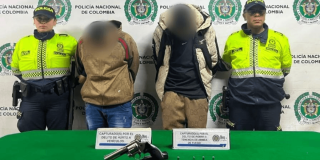 Dos capturados en medio de dos Policías. Sobre una mesa está el arma y los car