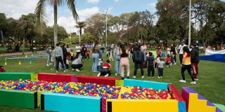 Personas adultas y menores participan en actividades recreativas sobre una zona verde de parque; en primer plano hay piscinas secas con pelotas de colores y al fondo se observan conos, árboles y grupos reunidos al aire libre.