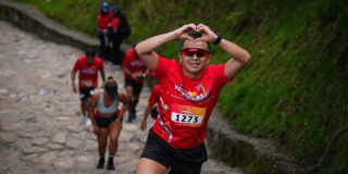Participantes ascienden por el sendero de Monserrate, en primer plano un corredor con el número 1273 realiza un gesto de corazón con las manos mientras continúa el recorrido.
