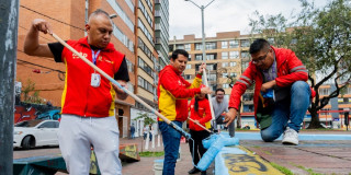 Grupo de trabajadores realiza labores de mantenimiento urbano pintando bordillos y superficies peatonales en espacio público, utilizando rodillos y pintura azul, con edificios residenciales y zona vial visibles al fondo.
