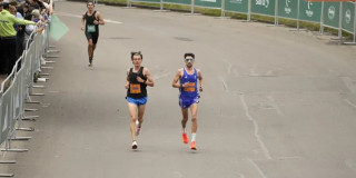 Grupo de atletas corre en la Media Maratón de Bogotá por una vía amplia durante competencia atlética urbana, con vallas metálicas a ambos lados y público observando desde el costado izquierdo