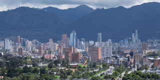Vista panorámica de Bogotá con edificios residenciales y empresariales, zonas verdes, la bandera de Colombia en primer plano y los cerros orientales bajo un cielo nublado.