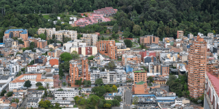 Vista panorámica de Bogotá con edificios residenciales y comerciales de diferentes alturas, calles arboladas y una extensa zona boscosa en los cerros orientales al fondo.