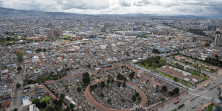 Vista aérea de Bogotá con extensa concentración de edificaciones, vías principales, cementerio central en primer plano y cielo cubierto de nubes sobre la ciudad.