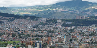 Panorámica de Bogotá con barrios densos, cerros verdes y cielo nublado fondo
