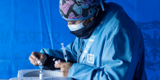 Imagen de una mujer vestida con chaqueta azul de la Secretaría de Salud preparando una jeringa para vacunar.