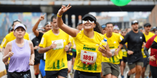 Corredores participan en la Media Maratón de Bogotá mientras avanzan por una avenida de la ciudad; en primer plano una atleta con camiseta amarilla y número 1891 saluda al público durante la competencia.