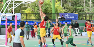 Jugadores disputan un balón bajo la canasta durante partido de baloncesto en los Juegos Intercolegiados, en una cancha cubierta con equipos identificados con uniformes rojos y verdes.