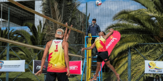 Partido de voleibol playa con dos deportistas junto a la red durante competencia al aire libre, con palmeras y juez en silla de arbitraje.