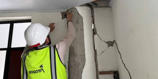 Persona con casco blanco y chaleco reflectivo con el logo de Bogotá inspecciona una columna y un muro interior con grietas visibles.