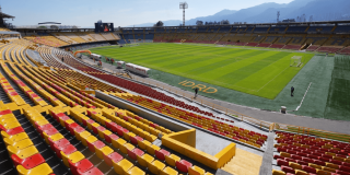 Vista interior del estadio El Campín en Bogotá con graderías amarillas y rojas, cancha de fútbol en óptimas condiciones y montañas al fondo.