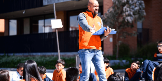 Foto que muestra un hombre con una carpeta en las manos y estudiantes sentados en el piso 