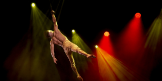 Imagen de un artista de circo en una puesta en escena al aire, de fondo se ven lices blancas y amarillas.