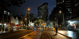 Ciclistas y peatones recorren una avenida iluminada durante la Ciclovía Nocturna de Bogotá, con edificios, comercio, alumbrado público y pavimento húmedo visibles en horario nocturno.