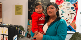 Una mujer con chaqueta verde sostiene en sus brazos a su bebé de 1 año que viste una sudadera roja