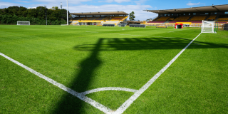 Cancha de fútbol del estadio de Techo con césped renovado, líneas demarcadas, arcos y graderías vacías bajo cielo parcialmente despejado.
