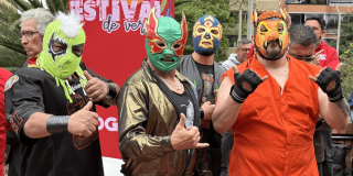 Cuatro luchadores con máscaras coloridas posan frente a un aviso del Festival de Verano durante una actividad de lucha libre en Bogotá.