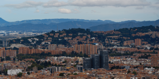 Vista panorámica de Bogotá con edificaciones residenciales y comerciales, zonas arborizadas y cerros al fondo bajo un cielo parcialmente nublado.