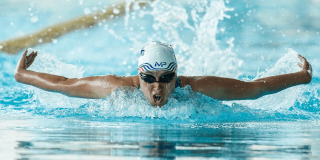 Persona practica natación estilo mariposa en una piscina de Bogotá, con gafas y gorro de natación mientras avanza entre salpicaduras de agua.