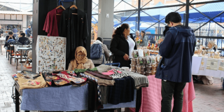 Foto que muestra un grupo de personas en una feria donde se exhiben artesanías y prendas de vestir.