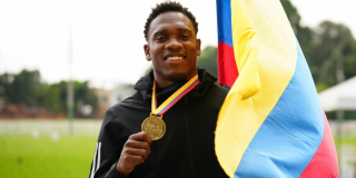 Ronal Longa sosteniendo una medalla dorada colgada al cuello y una bandera de Colombia. Al fondo se observa una pista o campo deportivo con vegetación desenfocada.