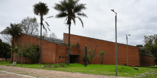 Fachada de ladrillo de una edificación rodeada de zonas verdes, con varias palmas y plantas ornamentales sembradas frente al muro.