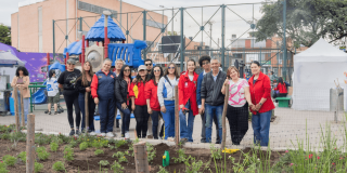 Grupo de personas con chaquetas rojas y ropa casual de pie junto a un jardín recién sembrado, frente a una estructura metálica y edificaciones del sector. En primer plano se observan plantas jóvenes y áreas con tierra.