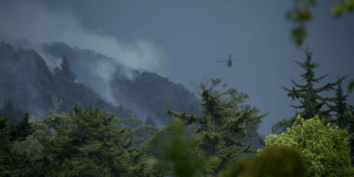Helicóptero con dispositivo para descarga de agua sobre una zona montañosa con presencia de humo, mientras se realizan labores de atención a un incendio forestal entre áreas boscosas.