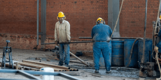 Obreros con cascos de seguridad trabajan en una obra de construcción, manipulando tuberías y equipos frente a una estructura de ladrillo.
