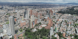 Vista aérea de un sector de Bogotá con edificios de gran altura, vías principales, zonas residenciales de alta densidad, áreas verdes y montañas al fondo bajo un cielo parcialmente nublado.