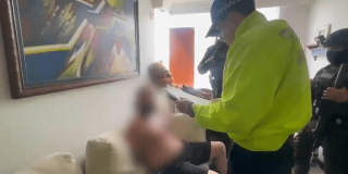 Persona con el rostro difuminado sentada en un sofá mientras otra persona con chaqueta reflectiva amarilla permanece de pie a su lado en una sala con cuadros en la pared
