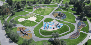 Vista aérea de un parque con amplias zonas verdes, senderos peatonales curvos y varias áreas de juegos infantiles distribuidas alrededor de una plazoleta central. Se observan estructuras de juego de diferentes tamaños, superficies de colores y árboles rodeando el espacio recreativo.