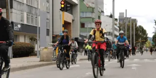Grupo de personas en bicicleta recorre una vía urbana durante una jornada de Ciclovía. En primer plano se observa una persona con casco y chaleco reflectivo, mientras detrás avanzan más ciclistas por una avenida rodeada de edificios y señalización vial.