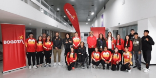 Grupo de deportistas del Equipo Bogotá, junto a personal de apoyo, posan para una fotografía en el hall interior de un edificio. Varias personas visten chaquetas deportivas en tonos rojo, amarillo y blanco.