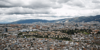 Vista panorámica de Bogotá con áreas residenciales y comerciales, edificios de diferentes alturas, vías urbanas y montañas al fondo bajo un cielo cubierto de nubes grises.